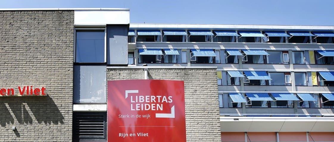 gevel met bord Libertas Leiden Rijn en Vliet, op de acchtergrond appartementencomplex van 4 of 5 verdiepeingen hoog, voor meerdere ramen uitgeklapte zonwering