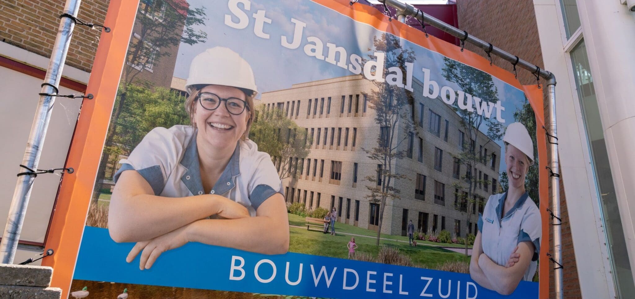 Grote bouwbanner met de tekst ‘St Jansdal bouwt – Bouwdeel Zuid’, met daarop twee glimlachende zorgmedewerkers met veiligheidshelmen voor een nieuw ziekenhuisgebouw in aanbouw