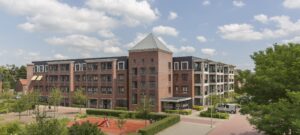 Foto van groot woongebouw, 4 verdiepingen, vooraan een toren, naar links en rechts woonvleugels. Op de voorgrond een speeltuintje en groen.