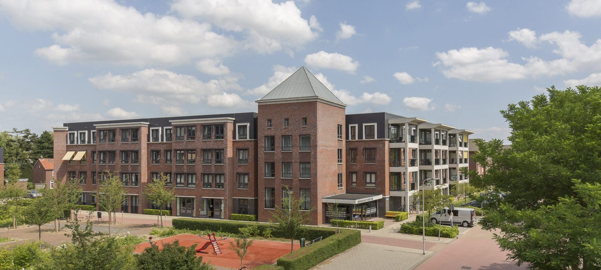Foto van groot woongebouw, 4 verdiepingen, vooraan een toren, naar links en rechts woonvleugels. Op de voorgrond een speeltuintje en groen.
