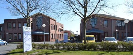 Twee identieke gebouwen met rode bakstenen gevels en blauwe raamaccenten. Op de voorgrond een wit informatiebord met tekst en logo’s langs een weg. Daarachter een parkeerplaats met enkele auto’s, afgescheiden door een lage heg en twee kale bomen. De ingang naar de parkeerplaats is zichtbaar achter het bord.