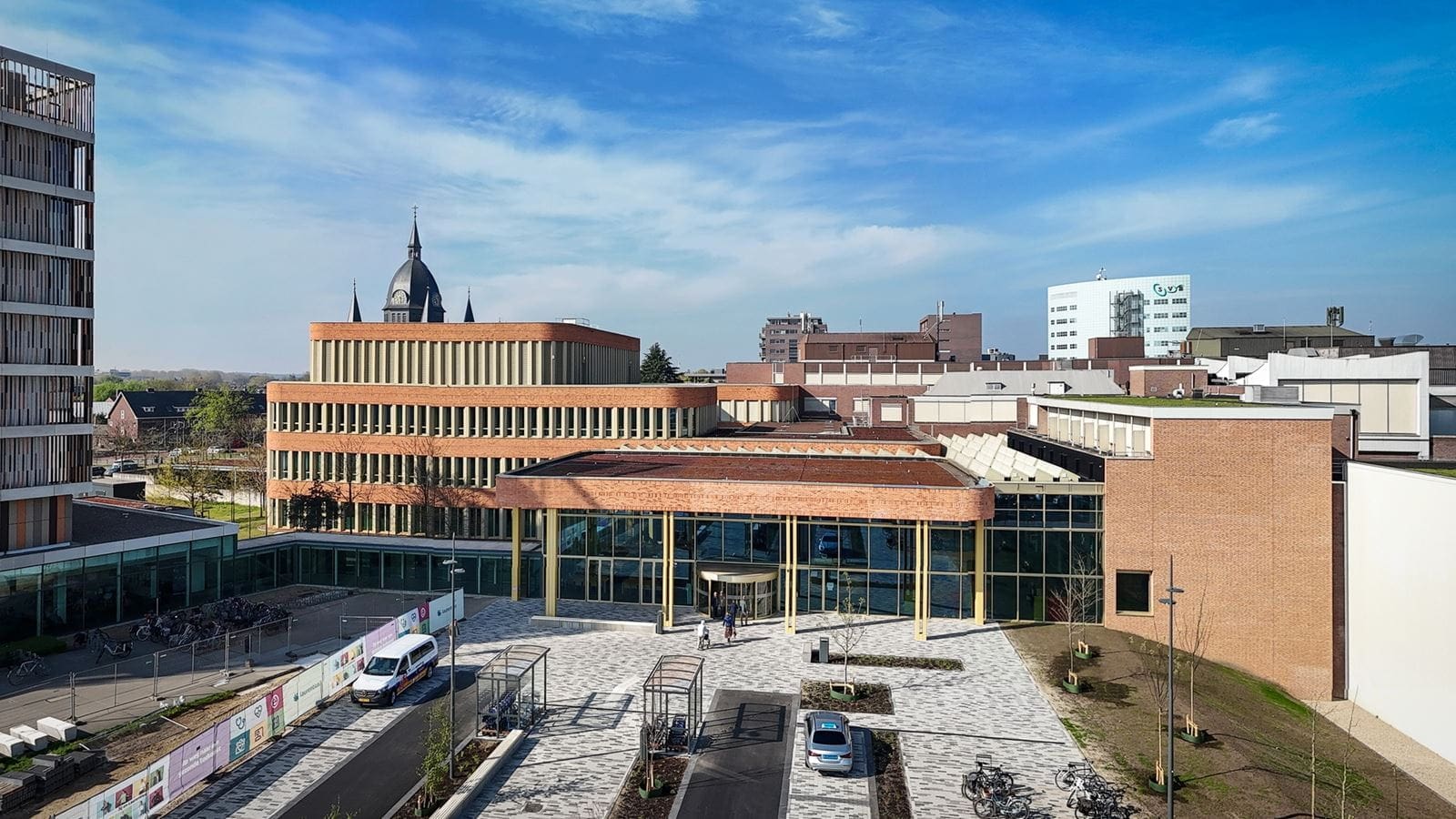 Een moderne, bakstenen gebouwencampus met glaspartijen en groene omgeving, centraal gelegen in een stedelijke omgeving met fietsen, auto’s en een historisch kerkgebouw op de achtergrond.