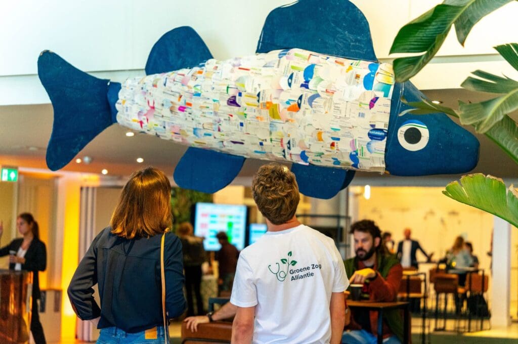 Een grote vis gemaakt van medicijnverpakkingen hangt boven bezoekers op een evenement, met logo "Groene Zog Alliantie" zichtbaar.