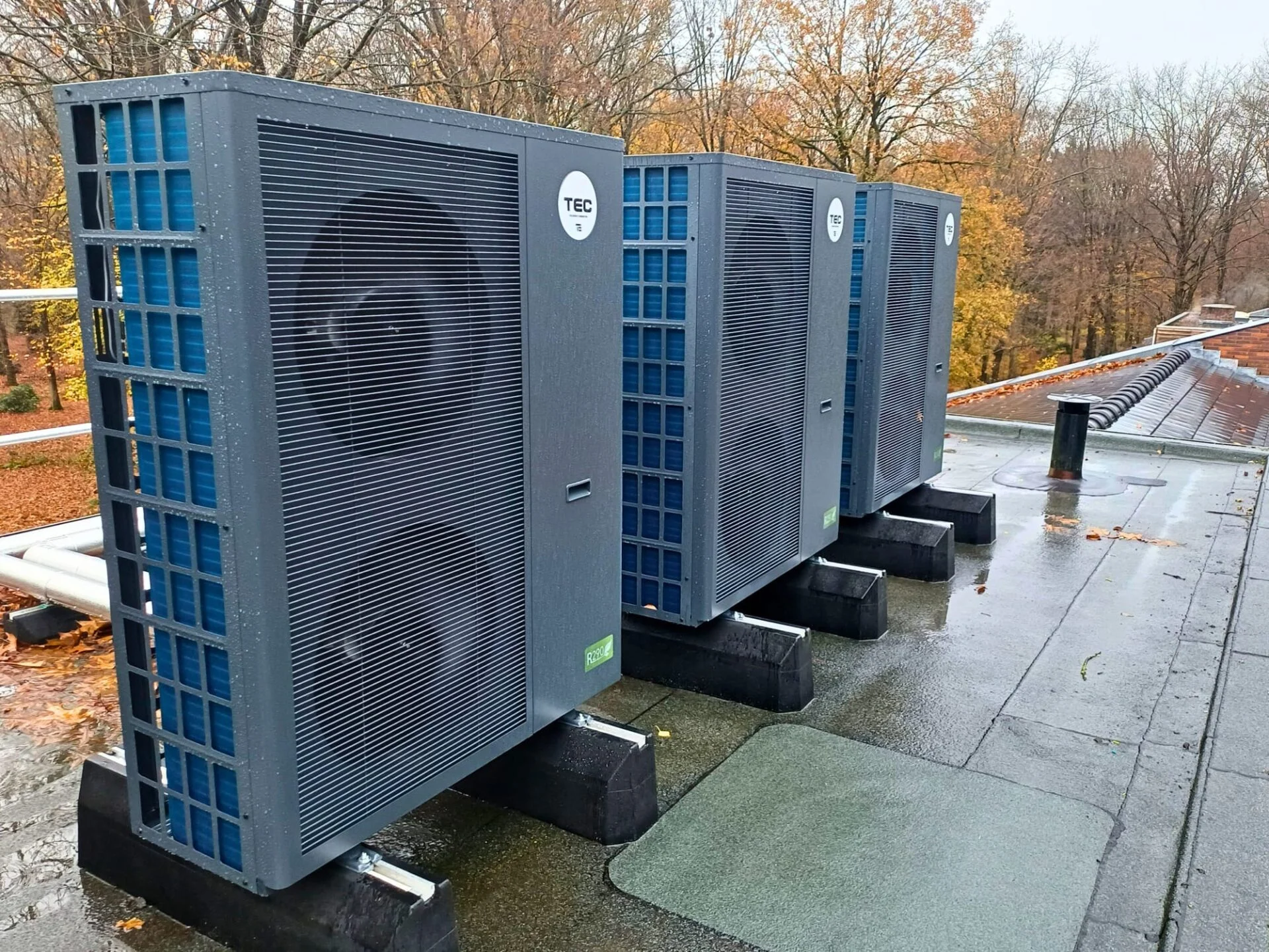 Drie warmtepompen van het merk TEC opgesteld op een plat dak, omgeven door herfstbomen op de achtergrond.