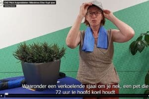 screenshot uit een video. iemand staat bij een tafeltje waarop een plant. Achtergrond is groene muur. Zet pet op hoofd en heeft blauw sjaaltje om nek