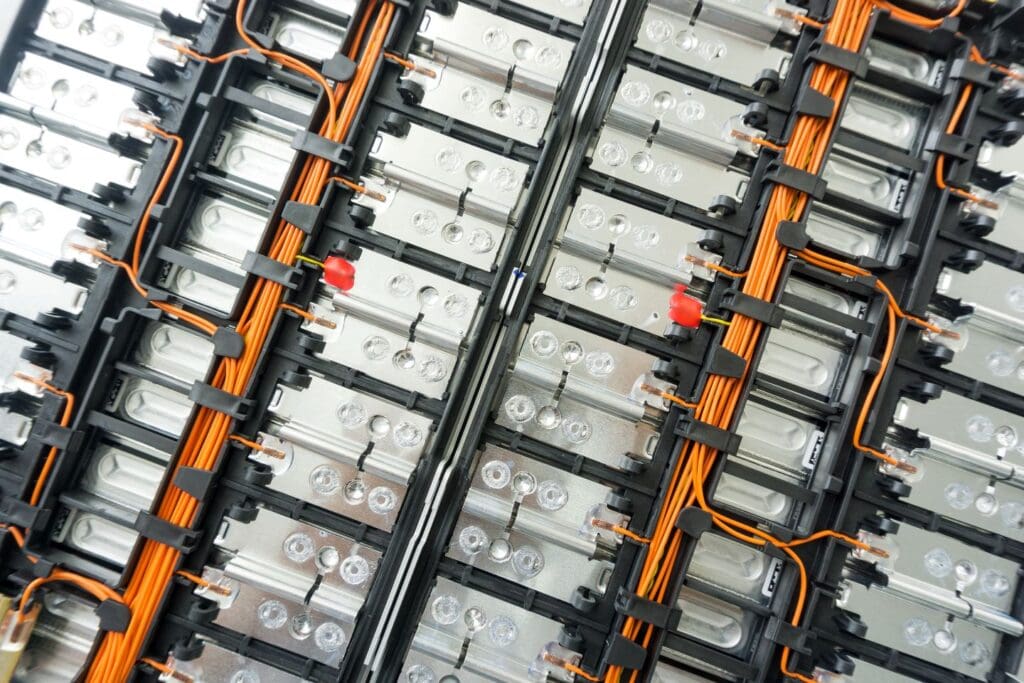 Dichte rij metalen batterijcellen met oranje kabels en rode connectoren, gemonteerd in een industriële opstelling.